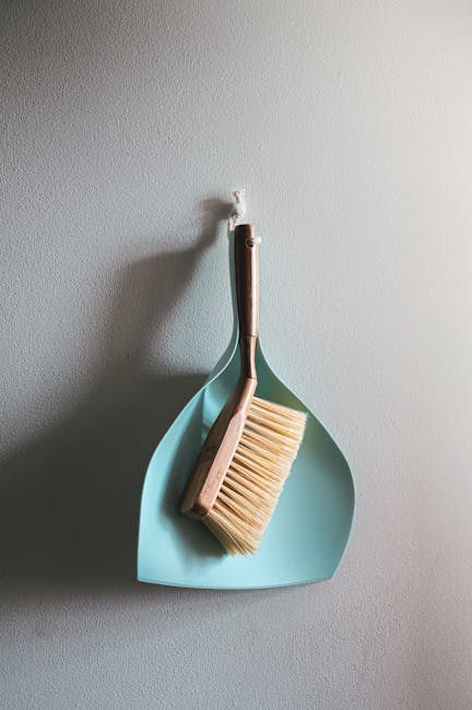 A light blue dustpan and a wooden-handled brush with beige bristles hanging on a plain, light-colored wall. The dustpan and brush are positioned with the brush resting inside the dustpan, which is hun